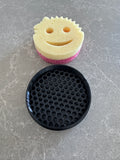 Smiley face yellow and pink kitchen sponge and black honeycomb-patterned soap saver on gray countertop