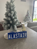 Decorative Christmas tree with a blue sign reading 'Alastair' on a textured wall. rego plate ppq