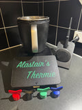 Black Thermomix mixer with silver handle, personalized mat reading 'Alastair's Thermie', colorful attachments on kitchen counter